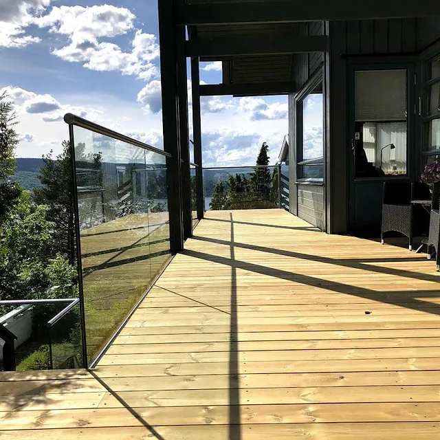 Terrasse med glassrekkverk og utsikt mot skog og åser under blå himmel.