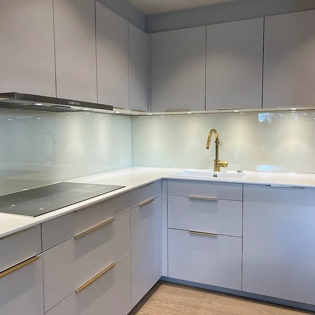 Moderne kjøkken med hvite skap, hvit benkeplate og glassplate på vegg bak vask og koketopp.